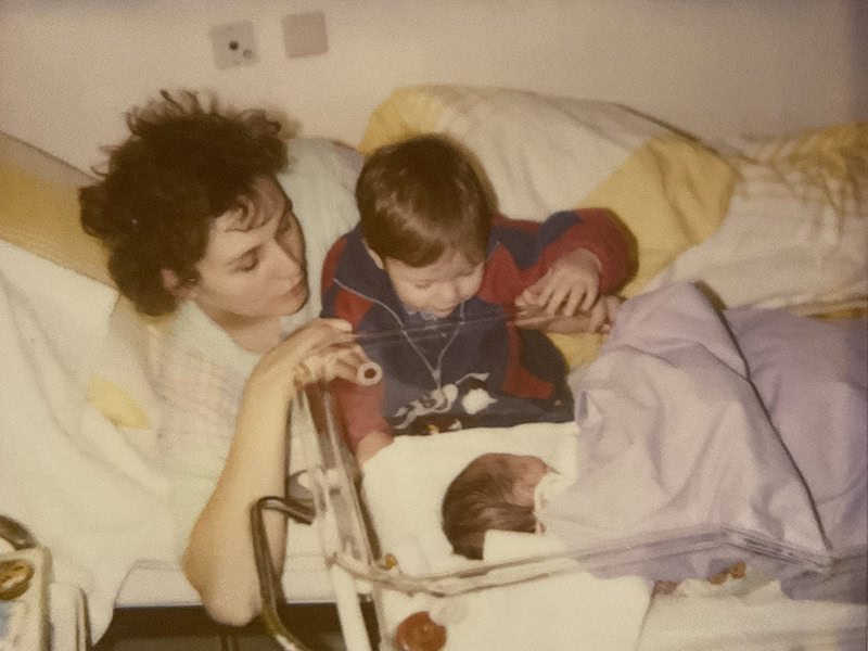 Schwangere Frau liegt im Krankenhausbett mit zwei Jungen, die ein Baby anschauen.