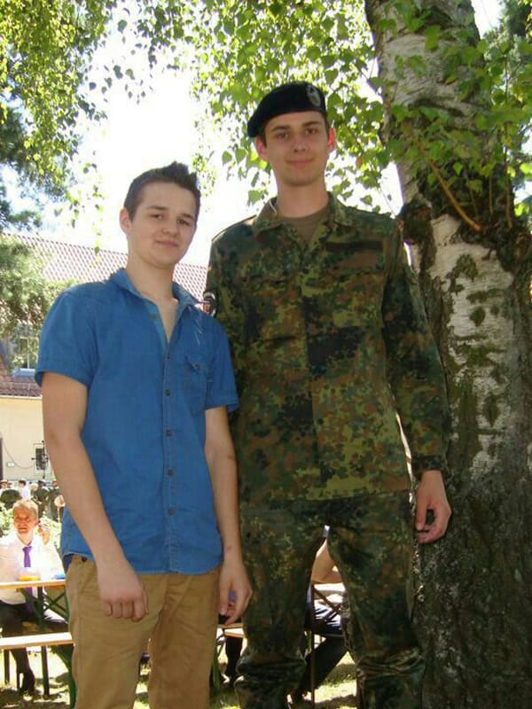 Junge im blauen Hemd und Soldat in Camouflage bei einem Outdoor-Event, lachend und freundlich, unter einer großen Baumkrone in sonnigem Garten.