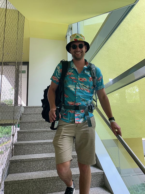 Fröhlicher Mann im Urlaubsoutfit mit Rucksack auf Treppe.