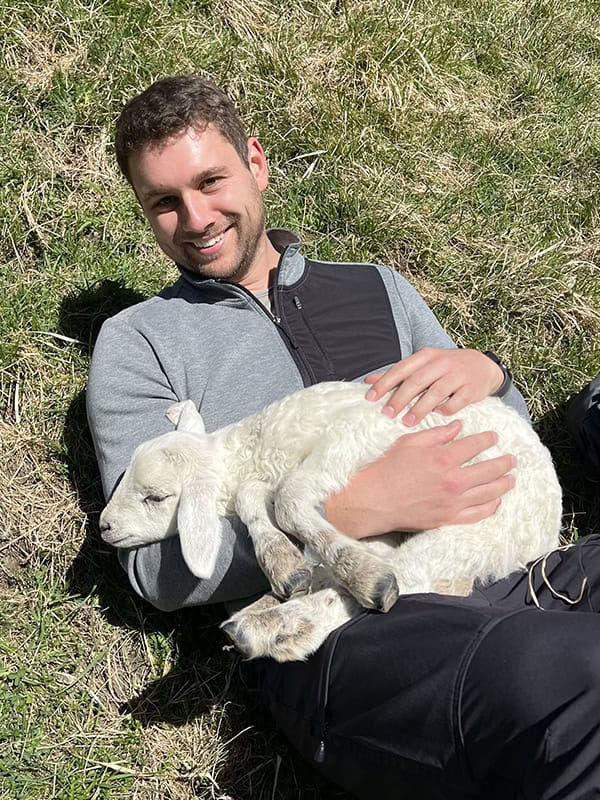 Junge Mann mit Ziegenbaby im Grünen, fröhliche Stimmung, Natur, Tierliebe, Geburtstag, Freude, ländliche Umgebung, Outdoor, Glücksmoment, Tierfreundschaft.