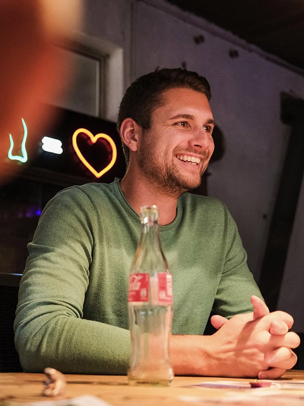 Freundlicher junger Mann beim Geburtstag, lächelnd mit Cola-Flasche, gemütliche Feieratmosphäre, Neonherz im Hintergrund, gemütliches Indoor-Event.