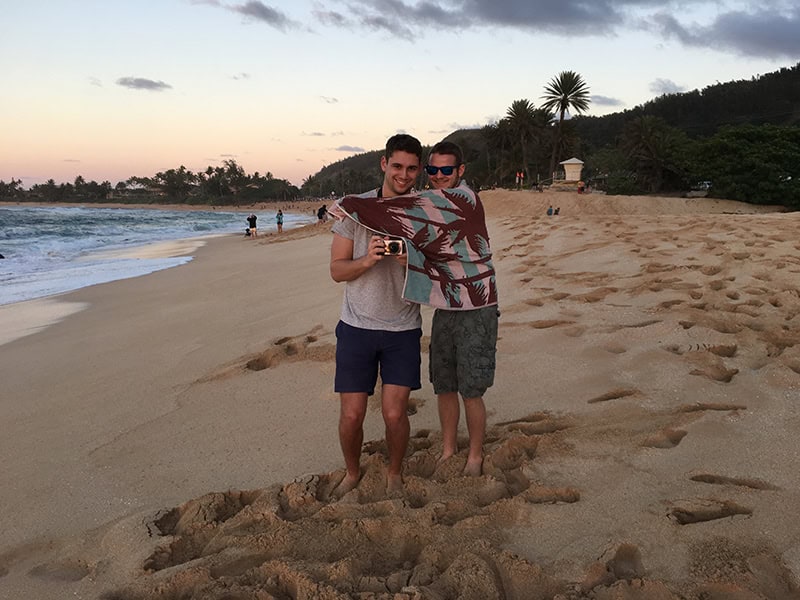 Fotografie von zwei Männern am Strand, einer hält eine Kamera, gemeinsames Lachen, entspannte Urlaubsstimmung, Sonnenuntergang, Palmen, tropisches Reiseziel.